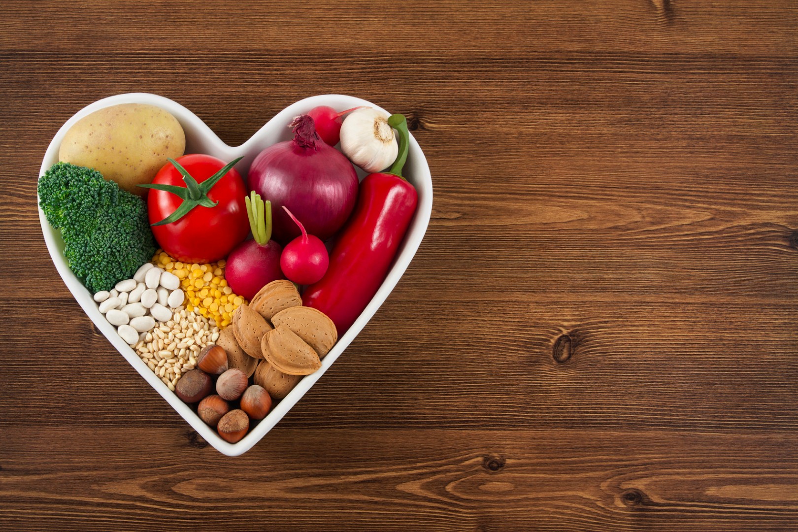 Heart health concept with related foods in white heart shaped bowl. potato red peppers, broccoli, radish, red onion, garlic, dry beans, almonds, nuts, and other pulses were arranged in heart space plate on white background.
