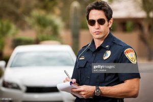 A mid-adult caucasian male police officer stands in front of a car, writing a ticket for a moving violation.