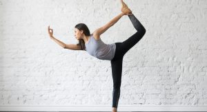 Young attractive woman practicing yoga, stretching in Natarajasana exercise, Lord of the Dance pose, working out, wearing sportswear, gray tank top, black pants, indoor full length, studio background