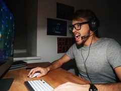 Excited handsome young gamer in glasses and earphones playing computer game and shouting at home