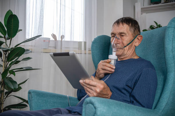 An elderly senior sits in a armchair with an oxygen mask and tablet in quarantine at home