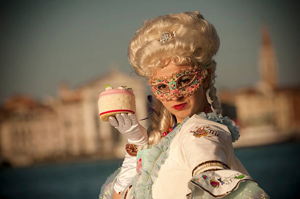 Beautiful young woman in attractive carnival costume with sweet cake in a hand posing on Isola di San Giorgio Maggiore. On a background we can see Piazza San Marco with Campanille. Venice, Italy.