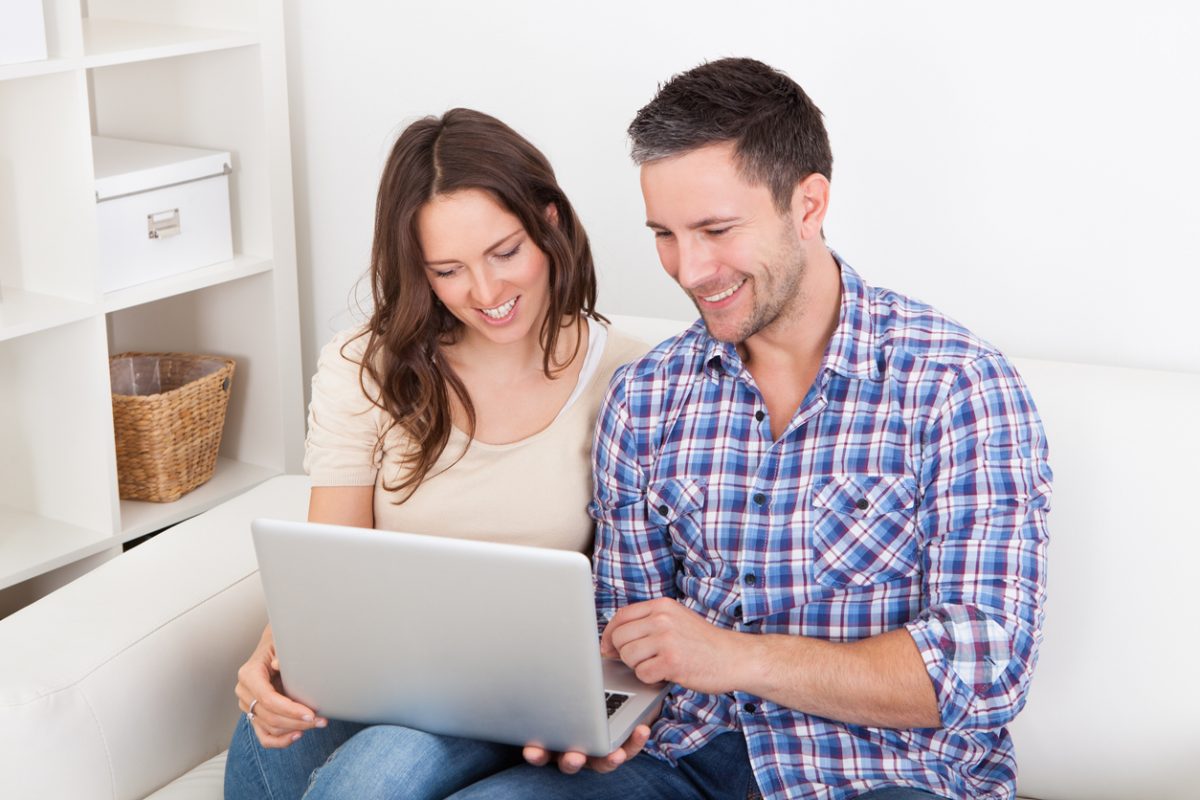 Portrait Of A Happy Young Couple Sitting On Couch Using Laptop