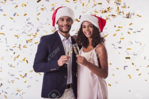 Elegant Afro American couple in Santa hats clinking glasses of champagne, looking at camera and smiling, on white background