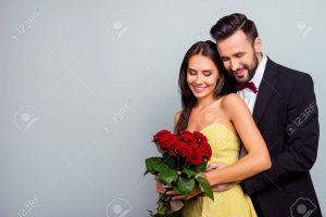 Portrait photo with copy space of charming, lovely, cute couple in formal wear, dress hugging and looking at bouquet of red roses, wife and husband celebrating 14 february on grey background